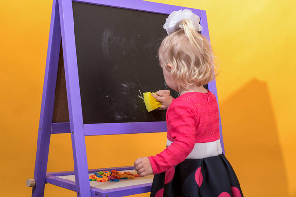 Petite fille essuie le tableau noir avec une éponge humide. Le concept d'éducation préscolaire pour les enfants. - Photo, image