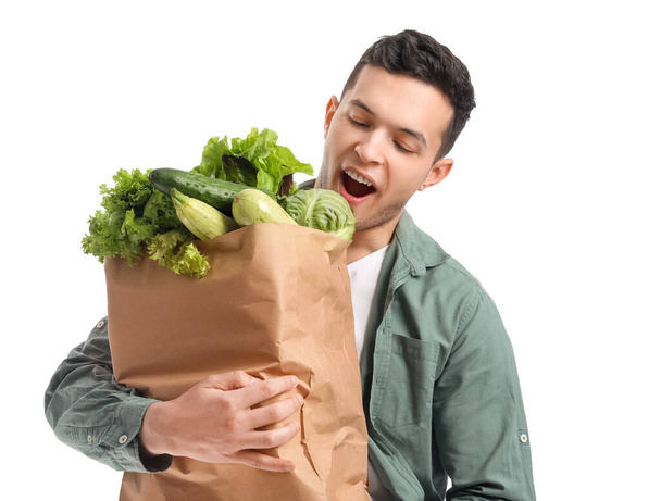 Удивленный молодой человек и бумажный пакет со свежими овощами на белом фоне - Фото, изображение