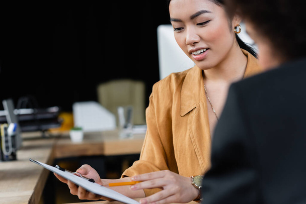asian businesswoman pointing with pencil at clipboard near blurred african american colleague - 写真・画像