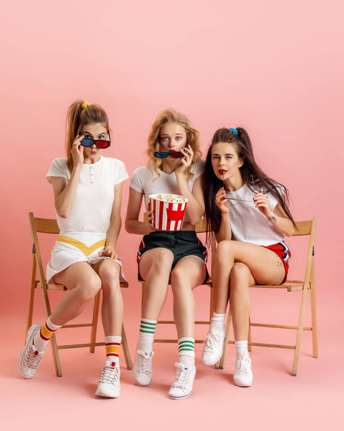 Three young women in retro 90s fashion style, outfits watching movie isolated over pink studio background. Concept of eras comparison, beauty, fashion and youth. - Valokuva, kuva