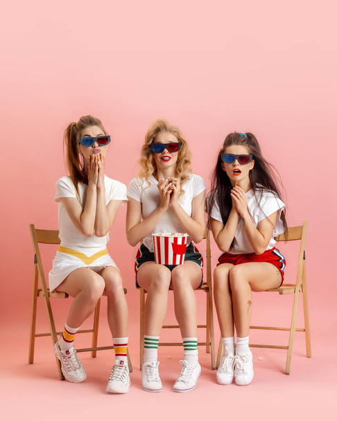 Three young women in retro 90s fashion style, outfits watching movie isolated over pink studio background. Concept of eras comparison, beauty, fashion and youth. - Fotó, kép