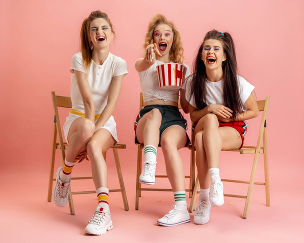 Three young women in retro 90s fashion style, outfits watching movie isolated over pink studio background. Concept of eras comparison, beauty, fashion and youth. - Foto, Bild