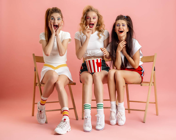 Three young women in retro 90s fashion style, outfits watching movie isolated over pink studio background. Concept of eras comparison, beauty, fashion and youth. - Фото, изображение