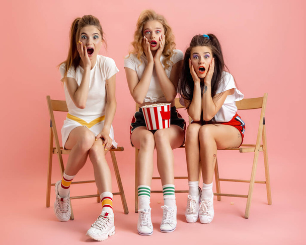 Three young women in retro 90s fashion style, outfits watching movie isolated over pink studio background. Concept of eras comparison, beauty, fashion and youth. - Фото, зображення