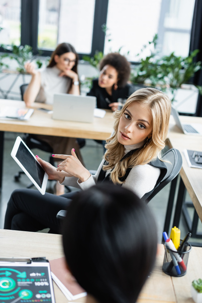 blonde woman pointing at digital tablet near blurred business partner in office - Foto, imagen