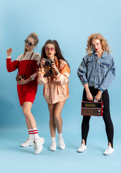 Pretty young women in retro 90s fashion style, outfits posing isolated over blue studio background. Concept of eras comparison, beauty, fashion and youth. - Foto, imagen