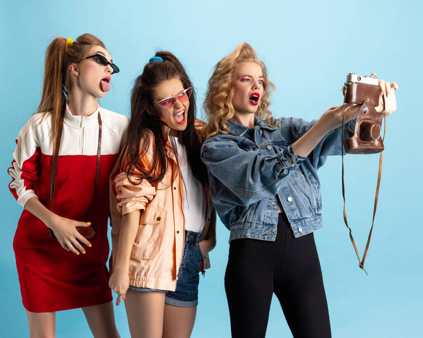 Three young girls in retro 90s fashion style, outfits posing isolated over blue studio background. Concept of eras comparison, beauty, fashion and youth. - Foto, afbeelding