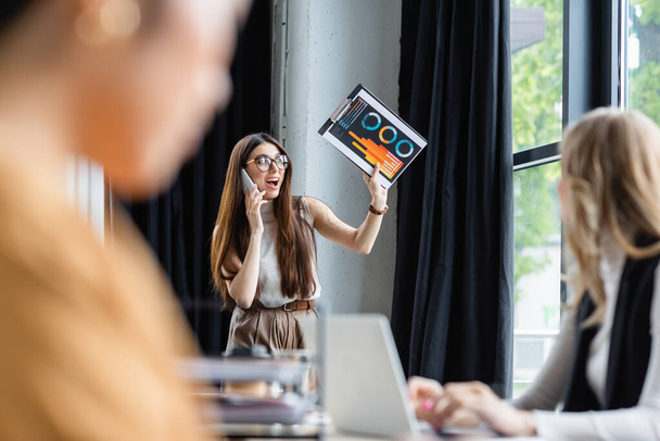 mulher de negócios animado acenando prancheta com gráficos enquanto conversa no smartphone perto de colegas embaçados - Foto, Imagem