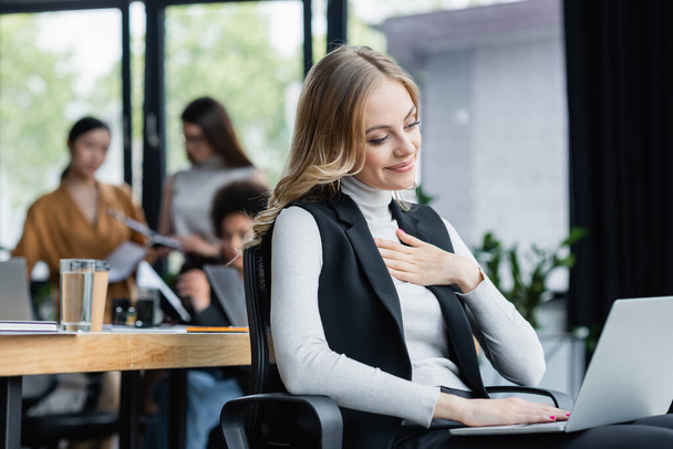 pleased businesswoman touching chest while working on laptop near blurred multicultural coworkers - Φωτογραφία, εικόνα