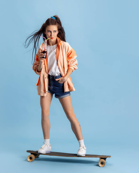 Beautiful adorable young girl in retro 90s fashion style, outfits on skateboard isolated over pink studio background. Concept of eras comparison, beauty, fashion and youth. - Foto, afbeelding