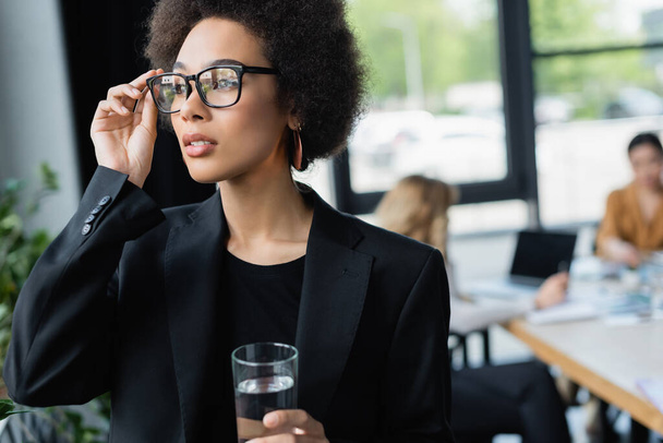 young african american businesswoman touching eyeglasses while standing with glass of water - Foto, imagen