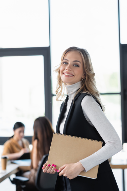 smiling businesswoman looking at camera while standing with paper folder in office - Photo, Image