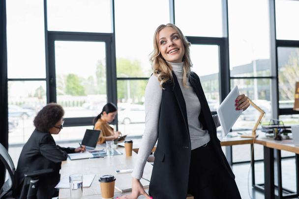 joyful businesswoman standing with digital tablet near blurred multiethnic colleagues in office - Foto, immagini