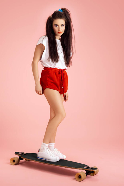 Beautiful adorable young girl in retro 90s fashion style, outfits on skateboard isolated over pink studio background. Concept of eras comparison, beauty, fashion and youth. - Фото, зображення