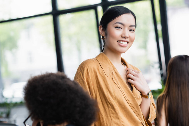 stylish asian businesswoman smiling at camera near blurred colleagues - Fotó, kép