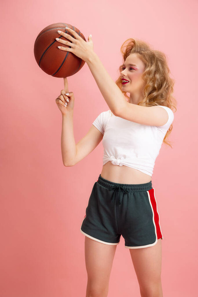 Portrait of young girl, tennis player in retro 90s fashion style, outfits playing with ball isolated over pink studio background. Concept of eras comparison, beauty, fashion and youth. - Fotografie, Obrázek