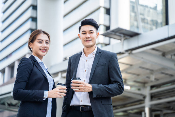 Азиатские молодые пары офисных деловых людей стоят на открытом воздухе в городе с уверенным лицом, держа горячий кофе. Рабочий мужчина и женщина чувствуют себя свежими и счастливыми с улыбкой после питья прохладительного напитка - Фото, изображение