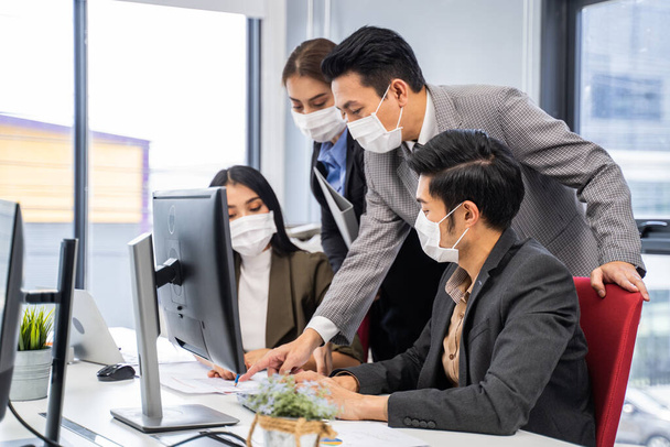 アジアの若いビジネスマンは、新しい通常のライフスタイルの概念でオフィスでコンピュータに取り組んでいます。人々は保護顔マスクを着用し、会社が再び再起動した後、 covidウイルスを防ぐために距離を保ちます. - 写真・画像