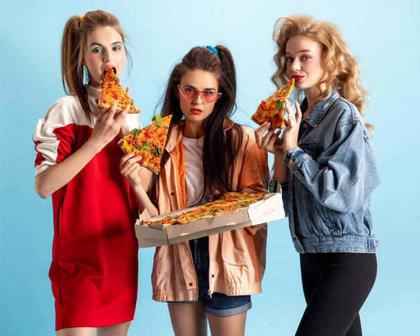 Three young attractive women in retro 90s fashion style, outfits eating pizza isolated over blue studio background. Concept of eras comparison, beauty, fashion and youth. - Fotó, kép