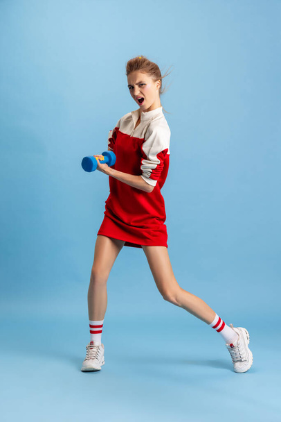 Portrait of young girl in retro 90s fashion style, outfits training with weights isolated over blue studio background. Concept of eras comparison, beauty, fashion and youth, humor - Foto, Bild