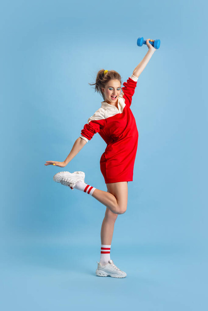 Portrait of young girl in retro 90s fashion style, outfits training with weights isolated over blue studio background. Concept of eras comparison, beauty, fashion and youth, humor - 写真・画像