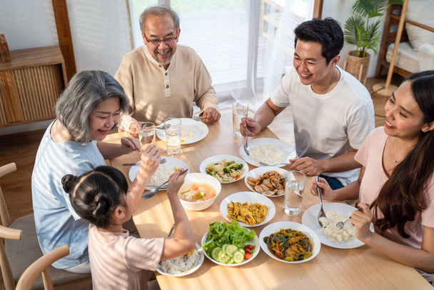 Ασιατική μεγάλη ευτυχισμένη οικογένεια περνούν χρόνο έχουν το μεσημεριανό γεύμα στο τραπέζι μαζί. Μικρή κόρη απολαμβάνει να τρώει τρόφιμα με τον πατέρα, τη μητέρα και τους παππούδες. Σχέση και δραστηριότητα πολλαπλών γενεών στο σπίτι - Φωτογραφία, εικόνα