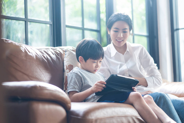 家庭学校アジアの小さな男の子は、コロナウイルスのパンデミックのためにデジタルリモートインターネット会議によって学校の先生からオンラインクラスを学ぶ。子供見タブレットコンピュータ上のソファとともに助けから母. - 写真・画像