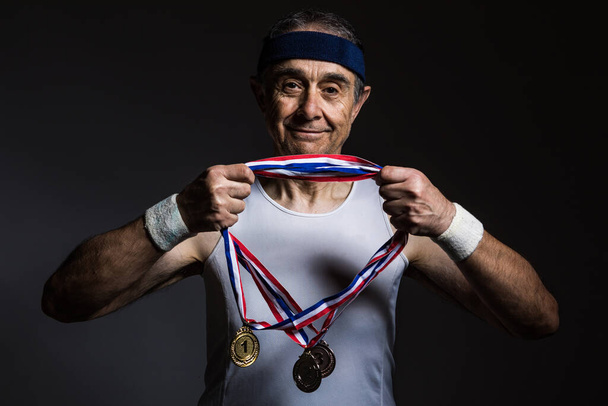 Un atleta anciano con una camisa blanca sin mangas, con marcas de sol en los brazos, tres medallas en el cuello, sobre un fondo oscuro. Concepto de deportes y victoria. - Foto, imagen