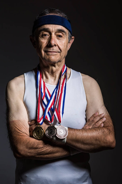 Anciano atleta con camisa blanca sin mangas, con marcas de sol en los brazos, con tres medallas en el cuello, con los brazos cruzados, sobre un fondo oscuro. Concepto de deportes y victoria. - Foto, imagen