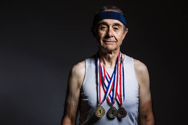 Atleta anciano con camiseta blanca, con marcas de sol en los brazos, con tres medallas en el cuello, mostrándolas, sobre un fondo oscuro. Concepto de deportes y victoria - Foto, imagen