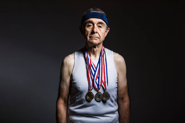 白いタンクトップを身に着けている高齢者のアスリート,腕の上に太陽のマークを持ちます,首に3つのメダルを持ちます,それらを示します,暗い背景に.スポーツと勝利のコンセプト - 写真・画像