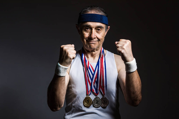 白い袖のないシャツを着て腕に太陽のマークがあり、首に3つのメダルが付いている高齢者のアスリートは、暗い背景で彼の拳をきれいにしています。スポーツと勝利のコンセプト. - 写真・画像