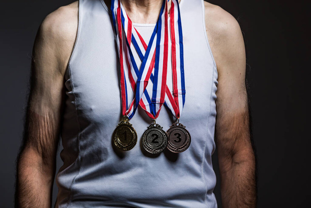 Elderly athlete wearing white tank top, with sun marks on the arms, with three medals on the neck, showing them, on a dark background. Sports and victory concept - Photo, Image