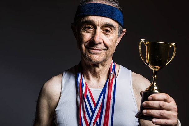Пожилой спортсмен в белой рубашке без рукавов, с пятнами от солнца на руках, с тремя медалями на шее, с трофеем в руках, на темном фоне. Концепция спорта и победы. - Фото, изображение