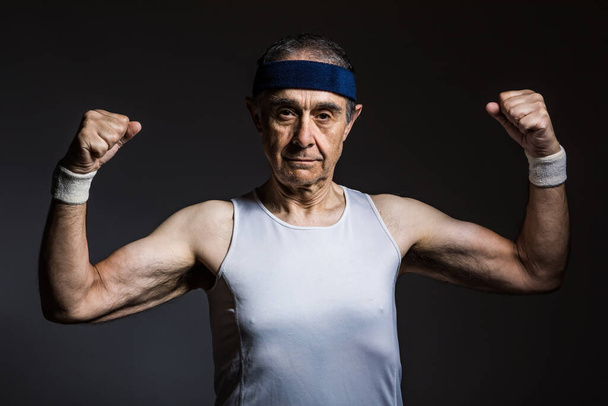 Elderly athlete wearing white sleeveless shirt, with sun marks on the arms, and blue headband, squeezing the biceps, on a dark background. Sports and victory concept. - Photo, Image