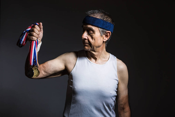 Un atleta anciano con una camisa blanca sin mangas, con marcas de sol en los brazos, con tres medallas en una de sus manos, sobre un fondo oscuro. Concepto de deportes y victoria. - Foto, imagen