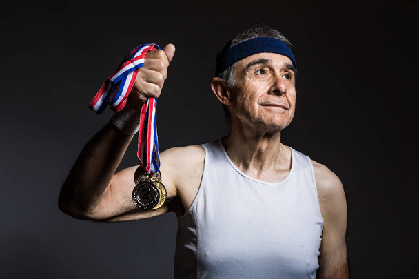 Elderly athlete wearing white sleeveless shirt, with sun marks on his arms, with three medals in one of his hands, on a dark background. Sports and victory concept. - Photo, Image
