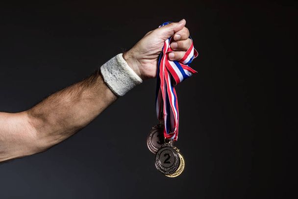 Arm of elderly athlete holding three gold, silver and bronze medals on a dark background. Sports and victory concept. - Photo, Image