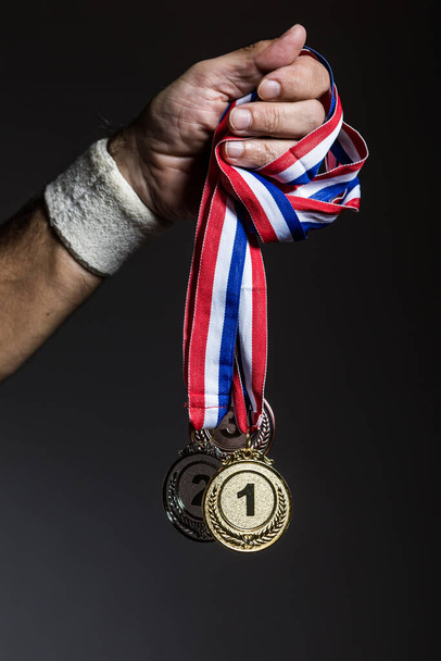 Arm of elderly athlete holding three gold, silver and bronze medals on a dark background. Sports and victory concept. - Photo, Image