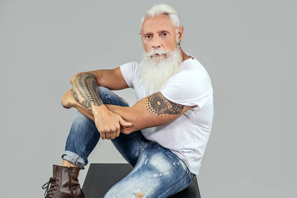 Fashionable senior man with white beard posing in studio, wearing white tshirt and jeans. Italian men with a lot of tattoos on his body.  - Foto, imagen