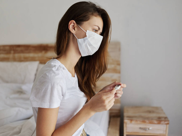 woman with thermometer in hands of medical mask bedroom checking temperature - Foto, Imagem