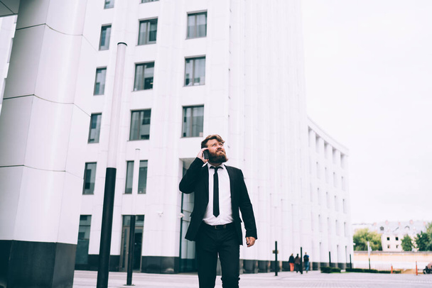 Serious bearded male with brown hair in black suit having conversation on mobile phone while walking near modern business buildings - Photo, Image
