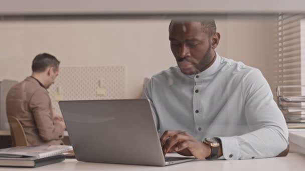 Střední detailní záběr mladého zamyšleného afroamerického muže v formální košili, sedícího za stolem v kanceláři, píšícího na přenosném počítači, pracujícího - Záběry, video