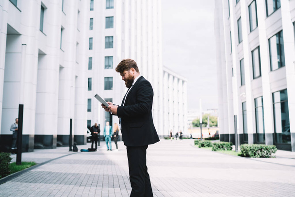 Side view of busy man looking at screen of gadget while standing on street against blurred exterior during successful conference - Фото, изображение