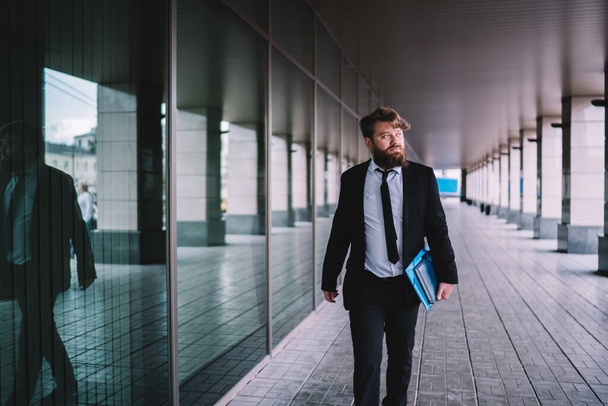 Thoughtful confident businessman with trendy haircut and stylish beard in formal outfit walking along glass wall with documents while looking away - Φωτογραφία, εικόνα