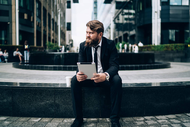 Serious bearded businessman in formal wear and tie sitting on stone border and looking away  thoughtfully while working on tablet - Fotografie, Obrázek