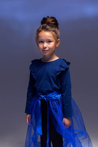 暗い背景に専門的にポーズをとる小さなモデルの女の子のファッショナブルな肖像画。黒のレギンスの女の子、青いジャケットと青い電車。子供服の概念 - 写真・画像