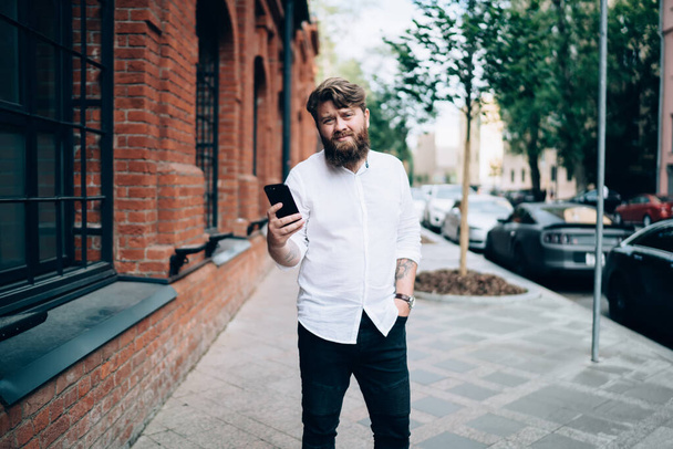 Happy bearded adult man in casual wear standing on street and chatting with friend on mobile phone while having break on daytime - Foto, Imagen
