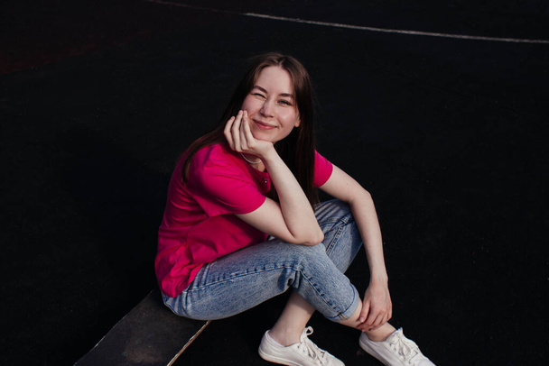 Smiling young woman in pink t-shirt winking and sitting on skateboard outside. Summer activity, healthy lifestyle, workout, sport, fitness, riding. Female skater. Having fun outdoors. - Photo, Image
