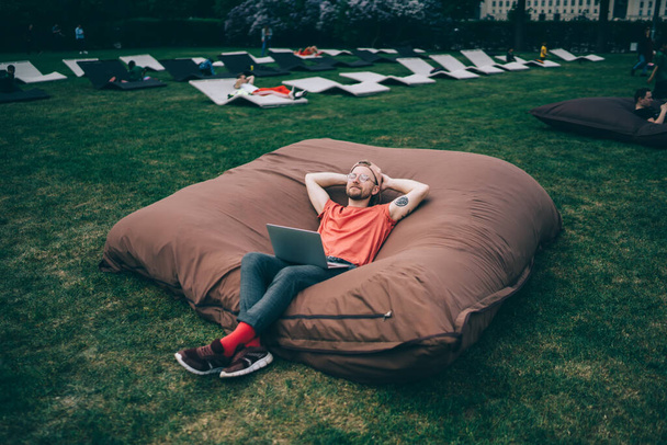 From above serene adult male in casual clothes with closed eyes chilling with hands behind head on big comfortable bean bag chair while using laptop on lawn - Foto, immagini